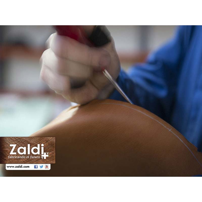 Mão segurando ferramenta sobre couro castanho com marcação branca, camisa azul, logotipo Zaldi e website.
