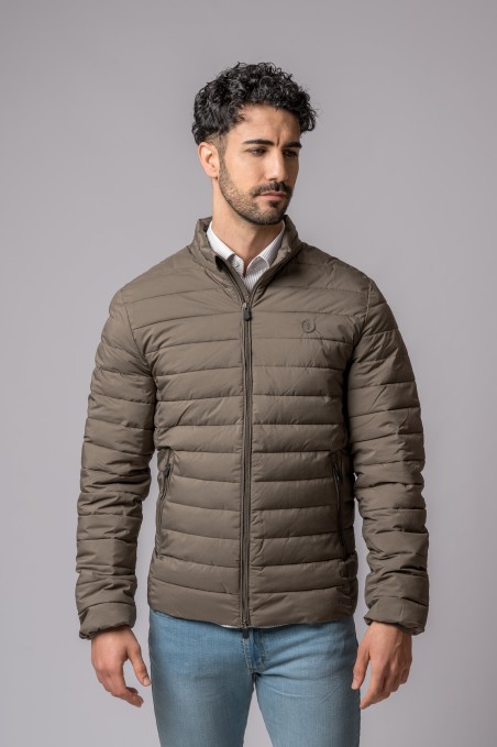 Homem com casaco acolchoado bege e calças azuis em fundo cinzento