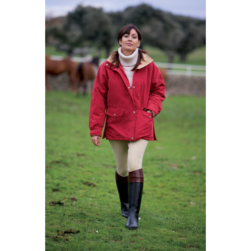 Mulher com casaco vermelho, calças bege e botas pretas a caminhar num campo com cavalos ao fundo.