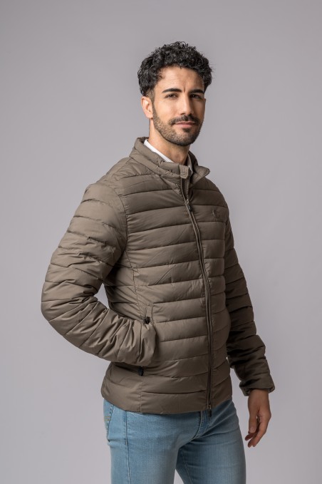 Homem a usar casaco acolchoado castanho com fecho de correr e calças de ganga, fundo cinzento