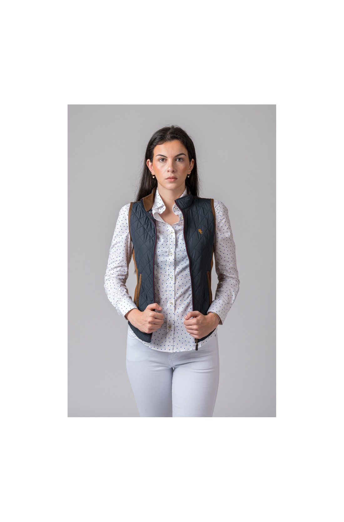 Mulher vestindo colete feminino azul escuro matelassê com detalhes castanhos e camisa branca com pontos, calças brancas, fundo cinzento.