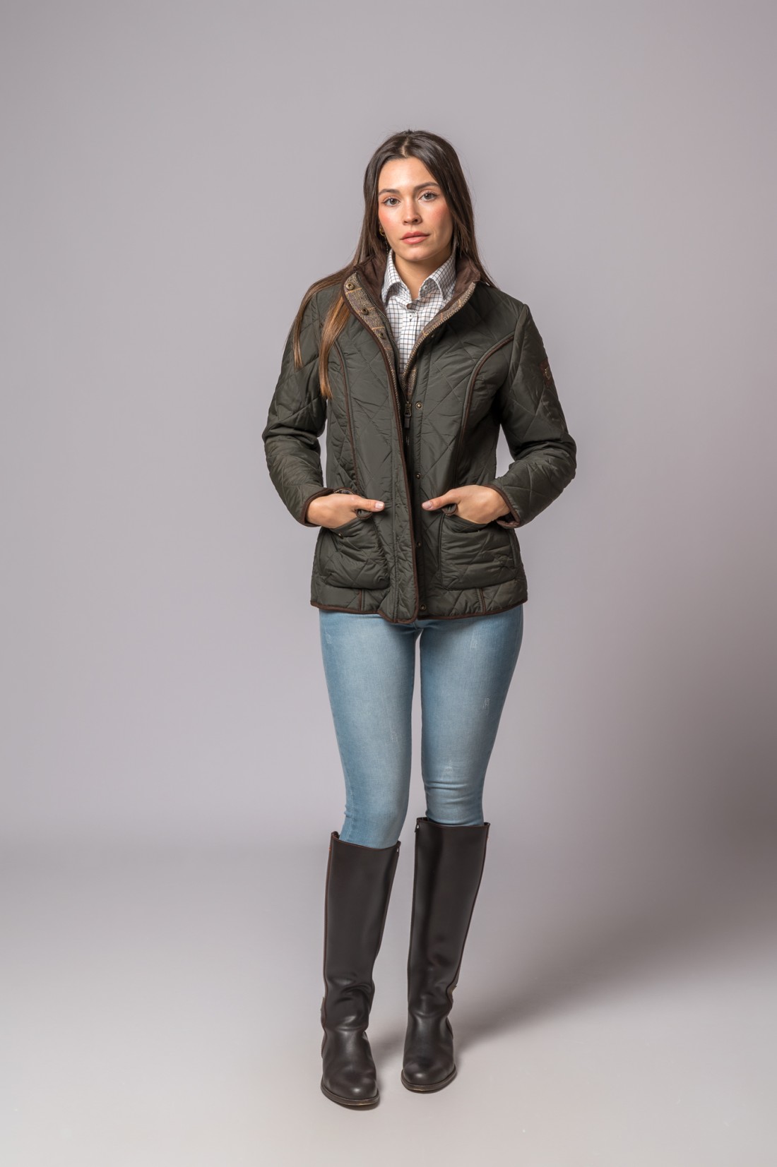 Mulher com casaco acolchoado verde, camisa quadriculada, jeans e botas castanhas, fundo cinza.