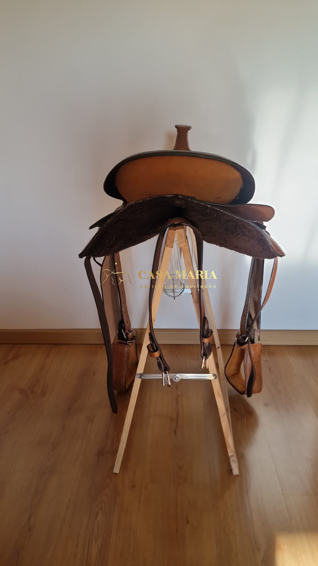 Sela de cavalo em couro castanho sobre estrutura branca no chão de madeira