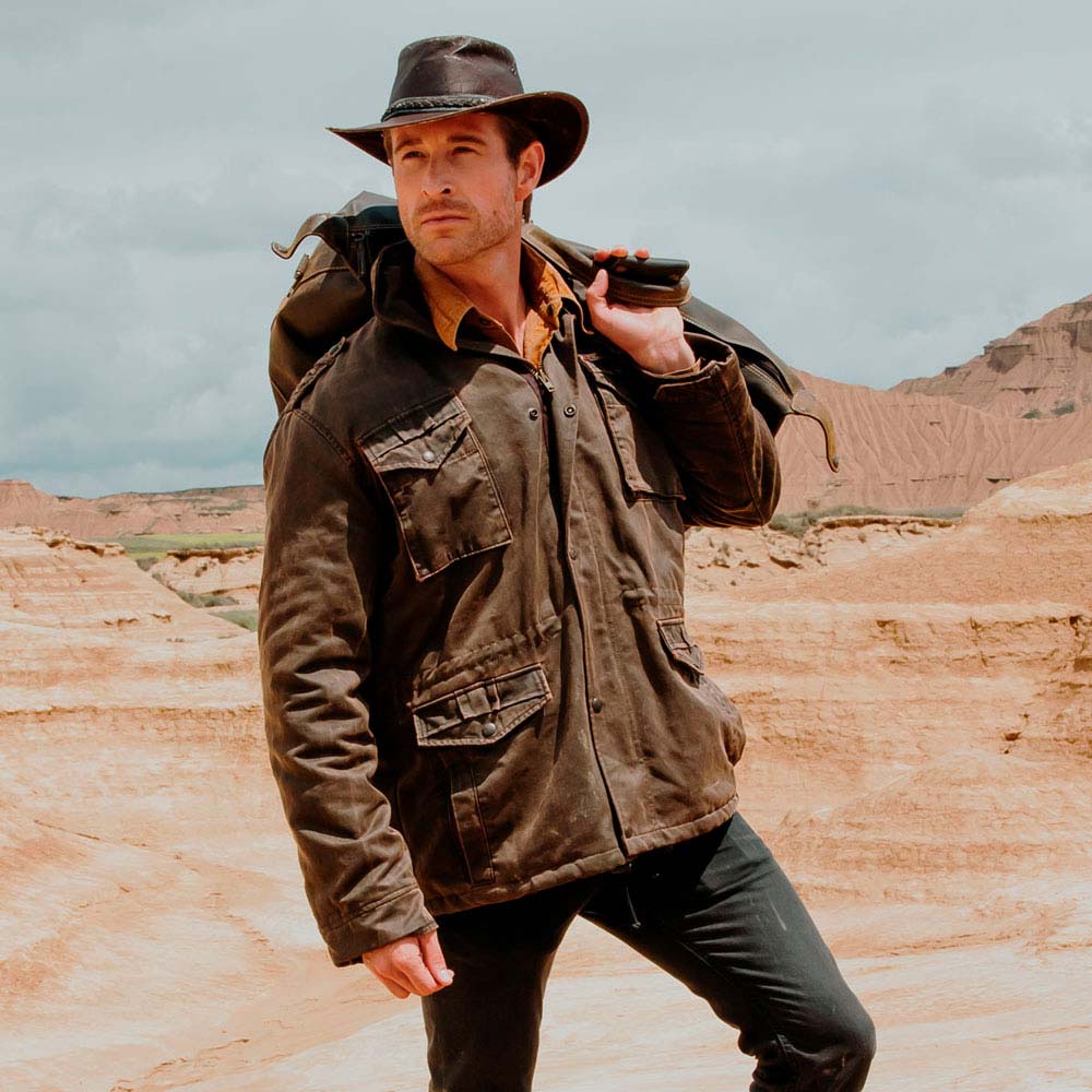 Homem com jaqueta castanha e chapéu, segurando mochila, em cenário desértico
