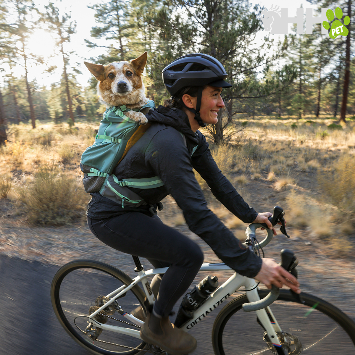 Pessoa de capacete com mochila verde transportando cão nas costas enquanto anda de bicicleta em floresta