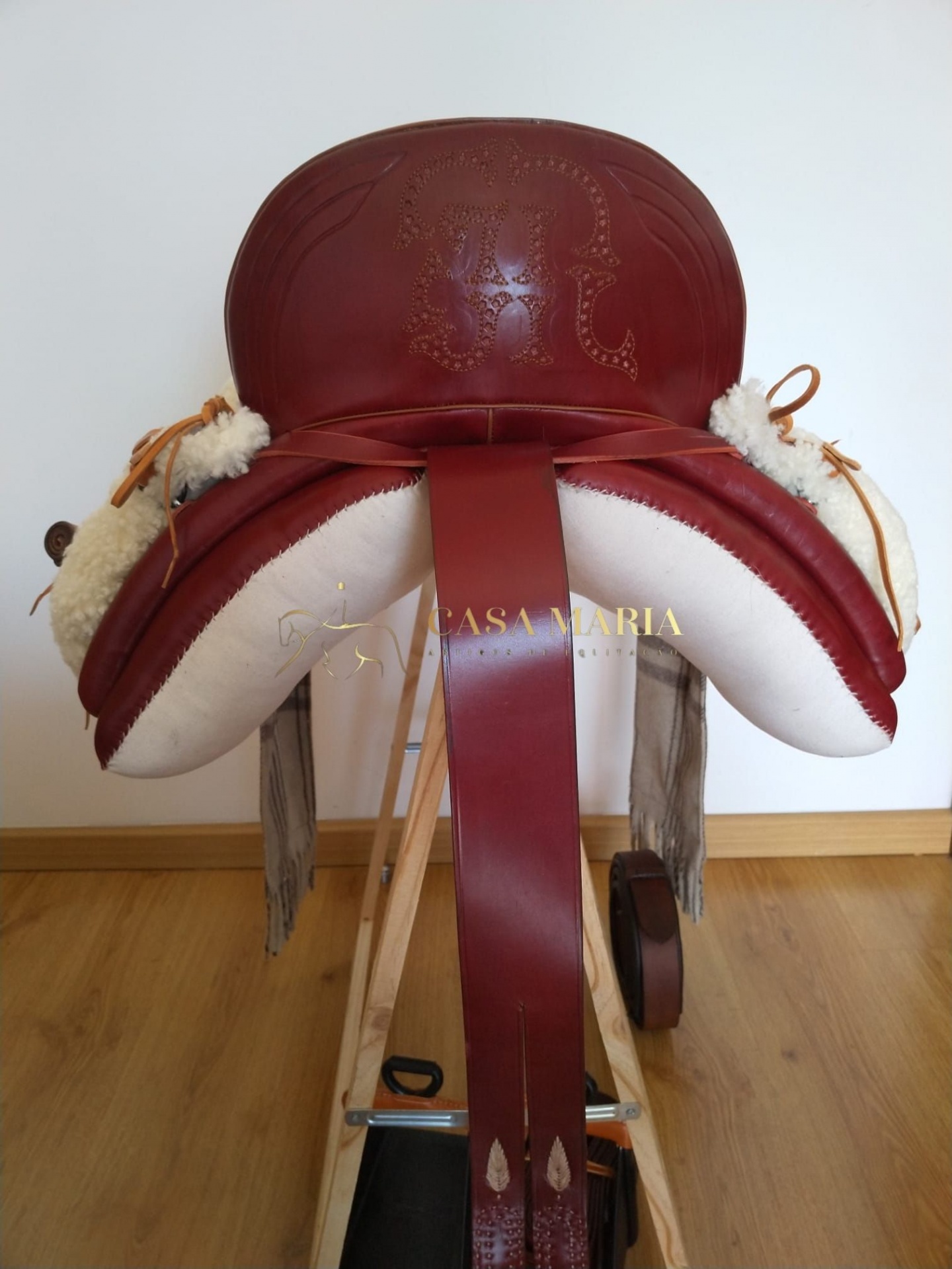 Sela de equitação vermelha e branca com detalhes em pele sintética branca sobre suporte de madeira.