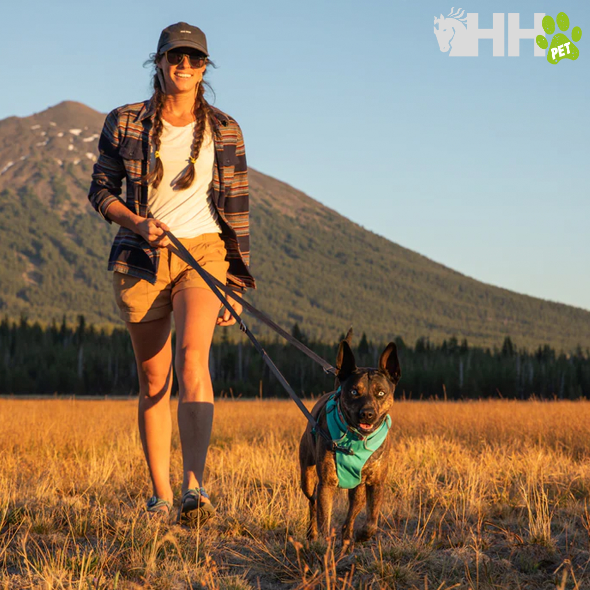 Mulher com boné e camisa às riscas a passear cão com bandana num campo aberto com montanha