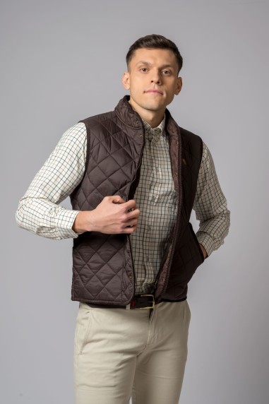 Colete masculino castanho escuro acolchoado sobre camisa de quadrados e calças claras.