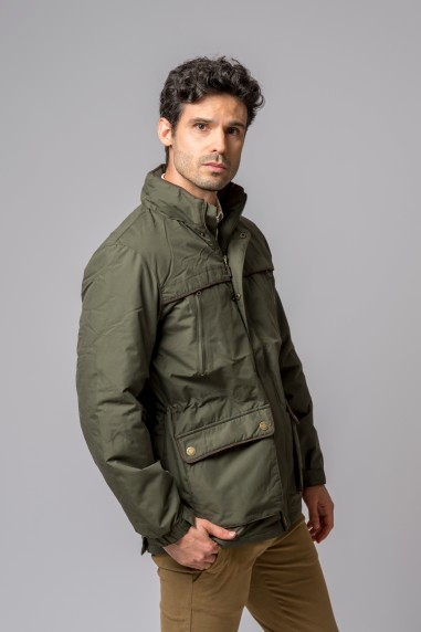 Homem de casaco verde-oliva com bolsos e calças castanhas em fundo cinzento