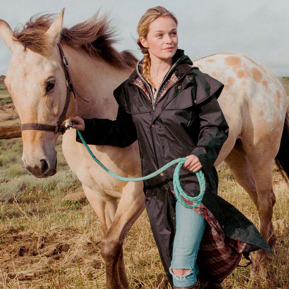 Mulher com casaco impermeável preto segurando corda ao lado de cavalo claro com manchas num campo