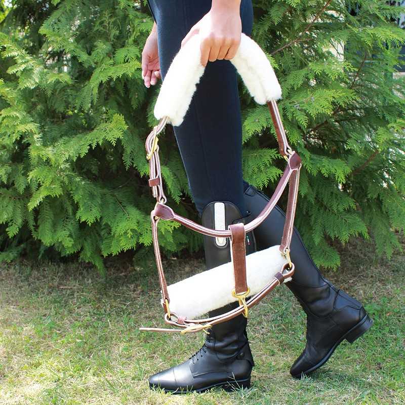 Pessoa a segurar rédea de equitação com acolchoamento branco e estrutura em couro castanho em ambiente exterior com relva e árvores verdes.