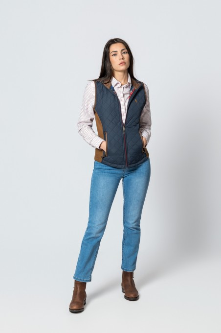 Mulher com colete acolchoado azul e detalhes castanhos, camisa xadrez, jeans e botas castanhas em fundo branco.