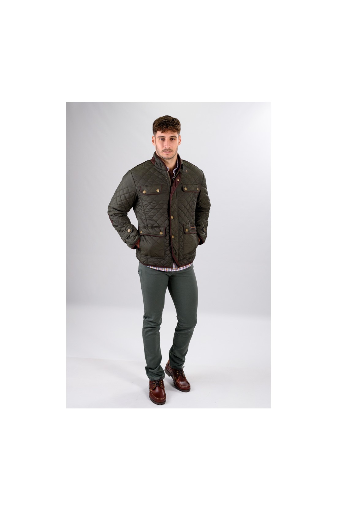 Homem com casaco matelassé verde-oliva, camisa aos quadrados, calças verdes e sapatos castanhos, fundo branco.