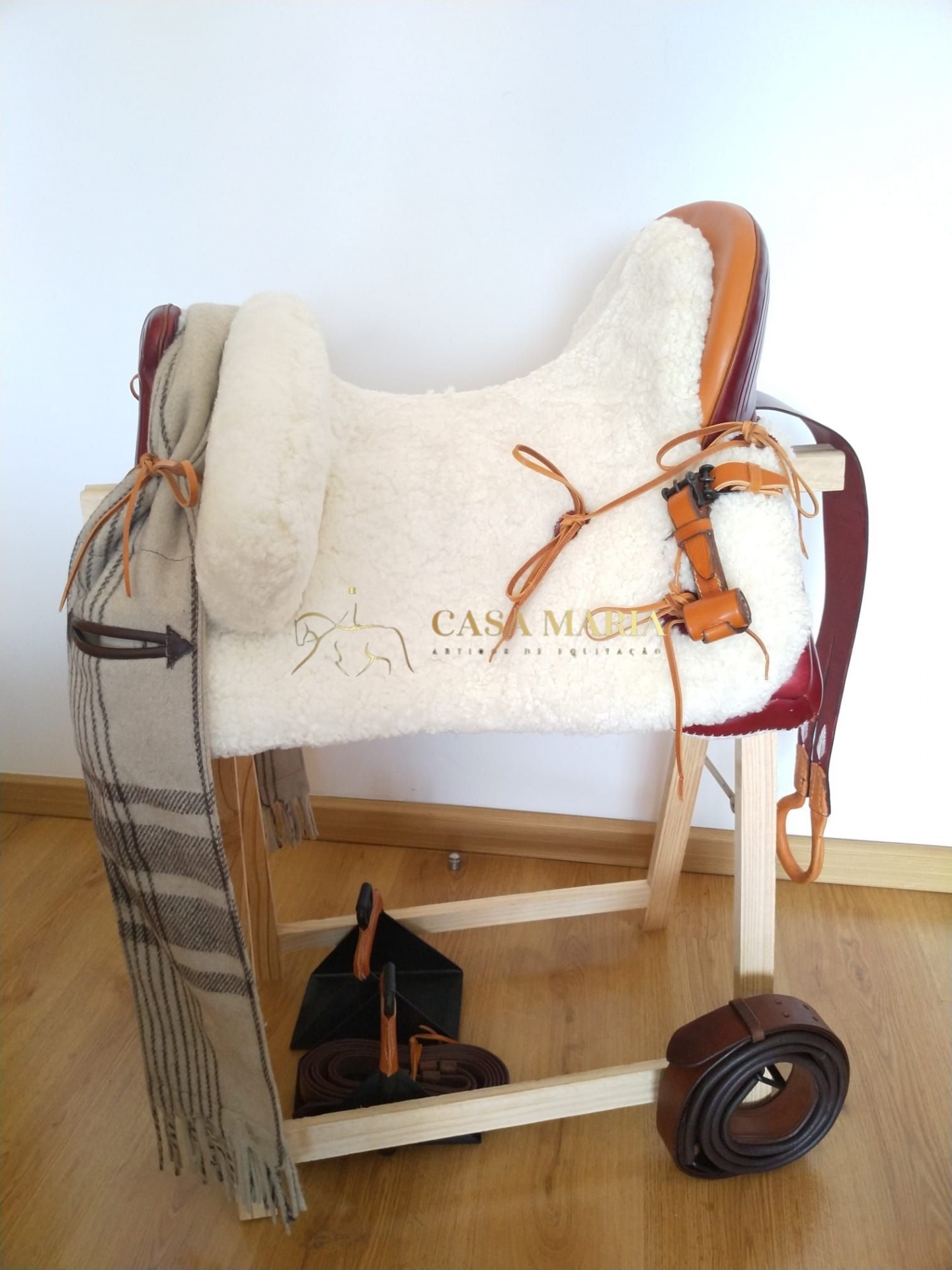 Sela de equitação em couro castanho com almofadado branco num suporte de madeira, junto com xale xadrez e arreios enrolados