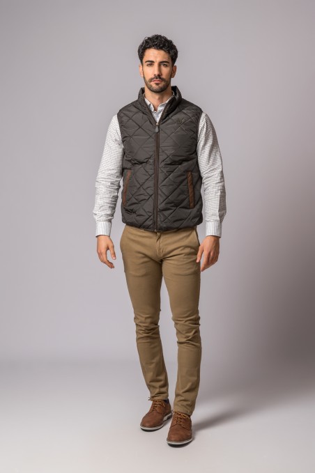 Homem vestindo colete acolchoado preto, camisa branca com padrão e calças bege