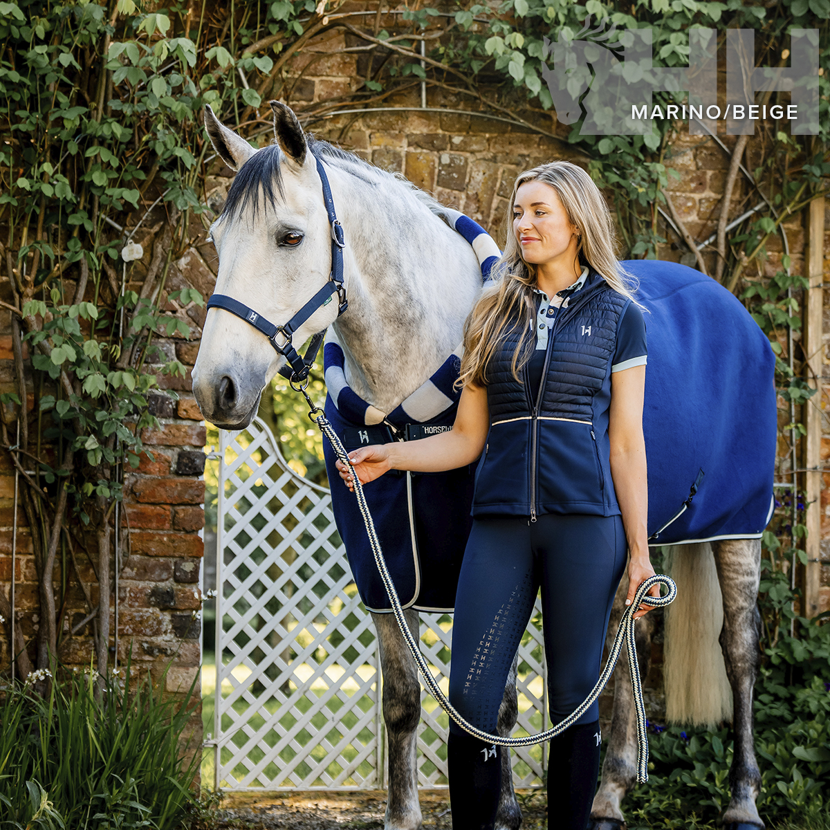 Mulher com roupa de equitação azul-marinho ao lado de cavalo branco com mantas e cabresto azul-marinho e bege perto de muro de tijolos e plantas