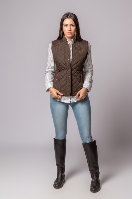 Mulher com colete castanho matelassê, camisa xadrez, jeans azuis e botas castanhas em fundo cinza