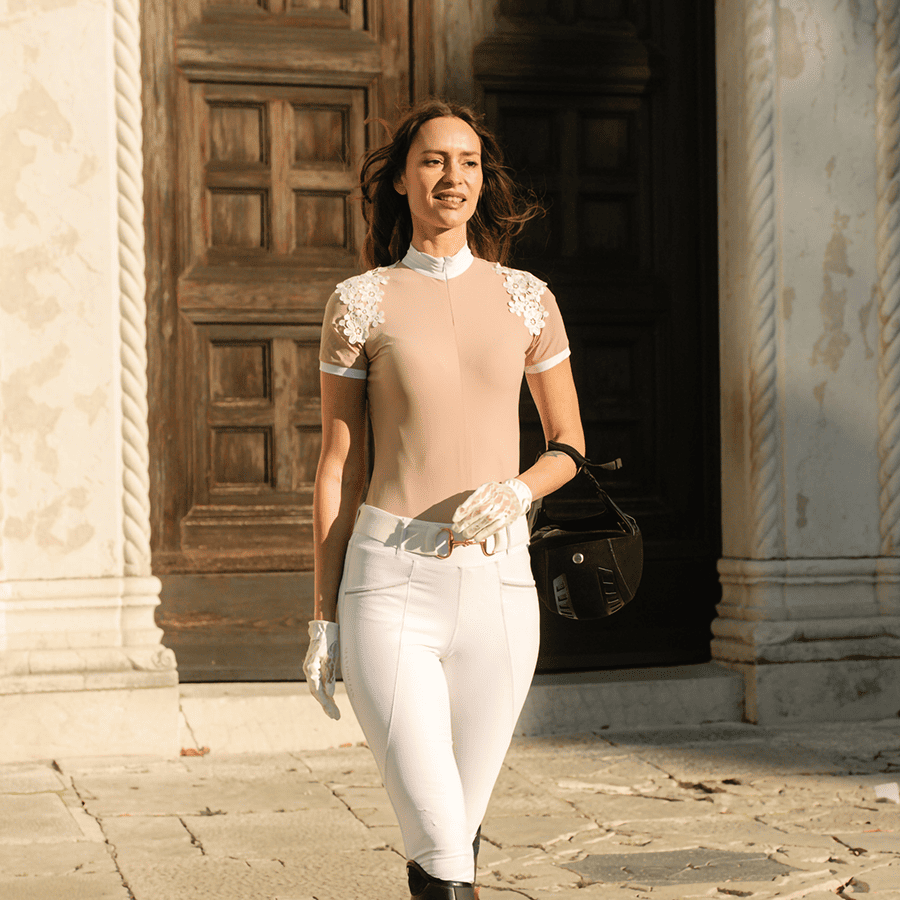 Mulher com roupa eqüestre bege e branca, luvas brancas e capacete preto numa rua com portas de madeira ao fundo.