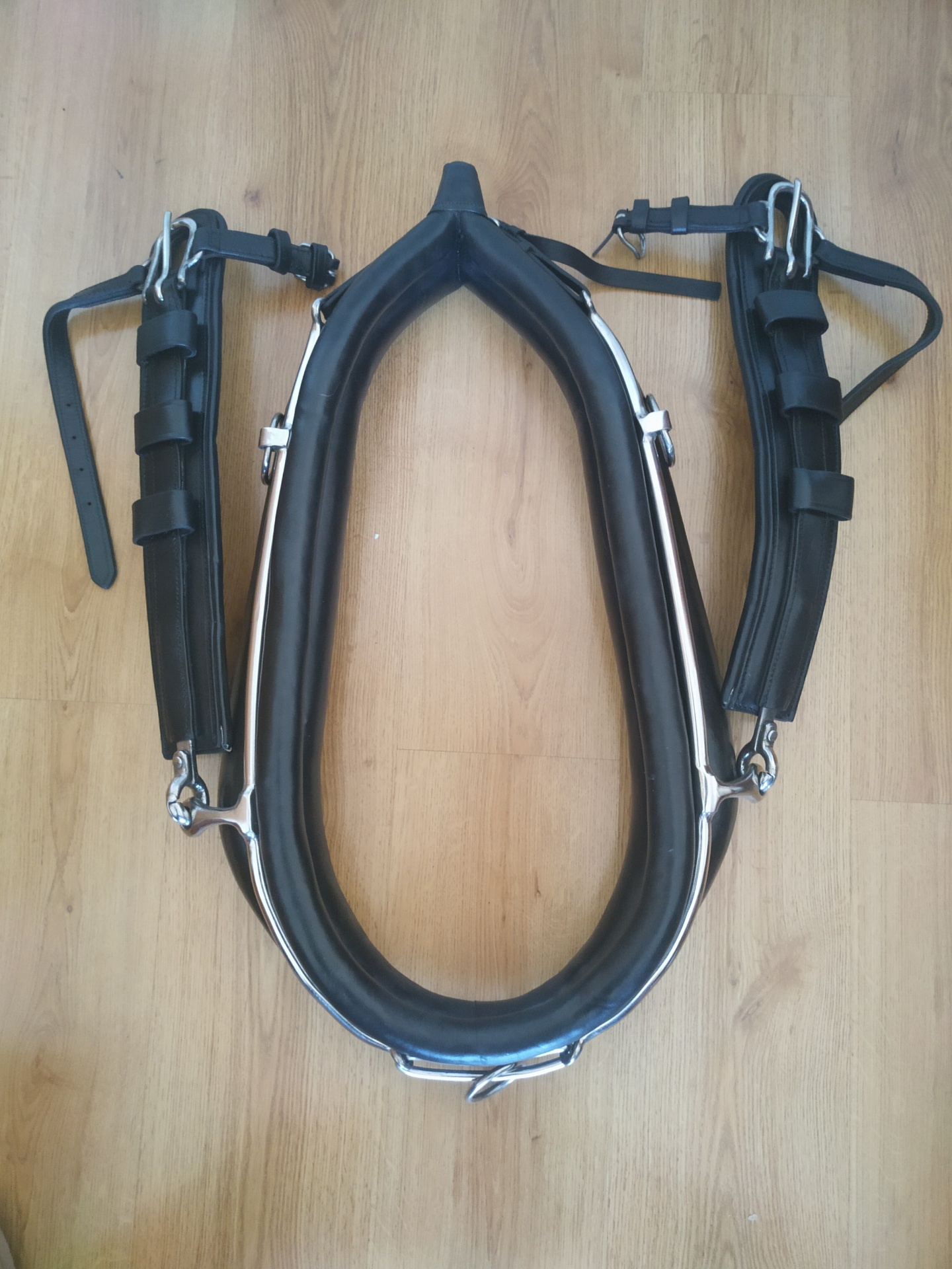 Arreio de cavalo preto almofadado com correias e fivelas num chão de madeira.