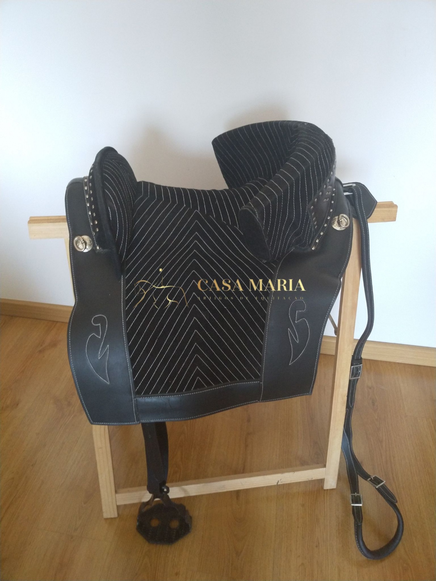 Sela preta de cavalo com costura branca e detalhes em couro pendurada em suporte de madeira