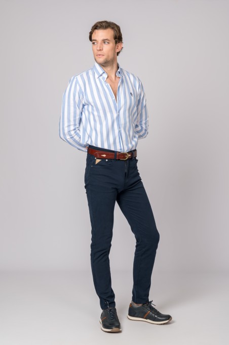 Homem com camisa azul e branca às riscas, calças azuis escuras, cinto castanho e sapatos desportivos pretos