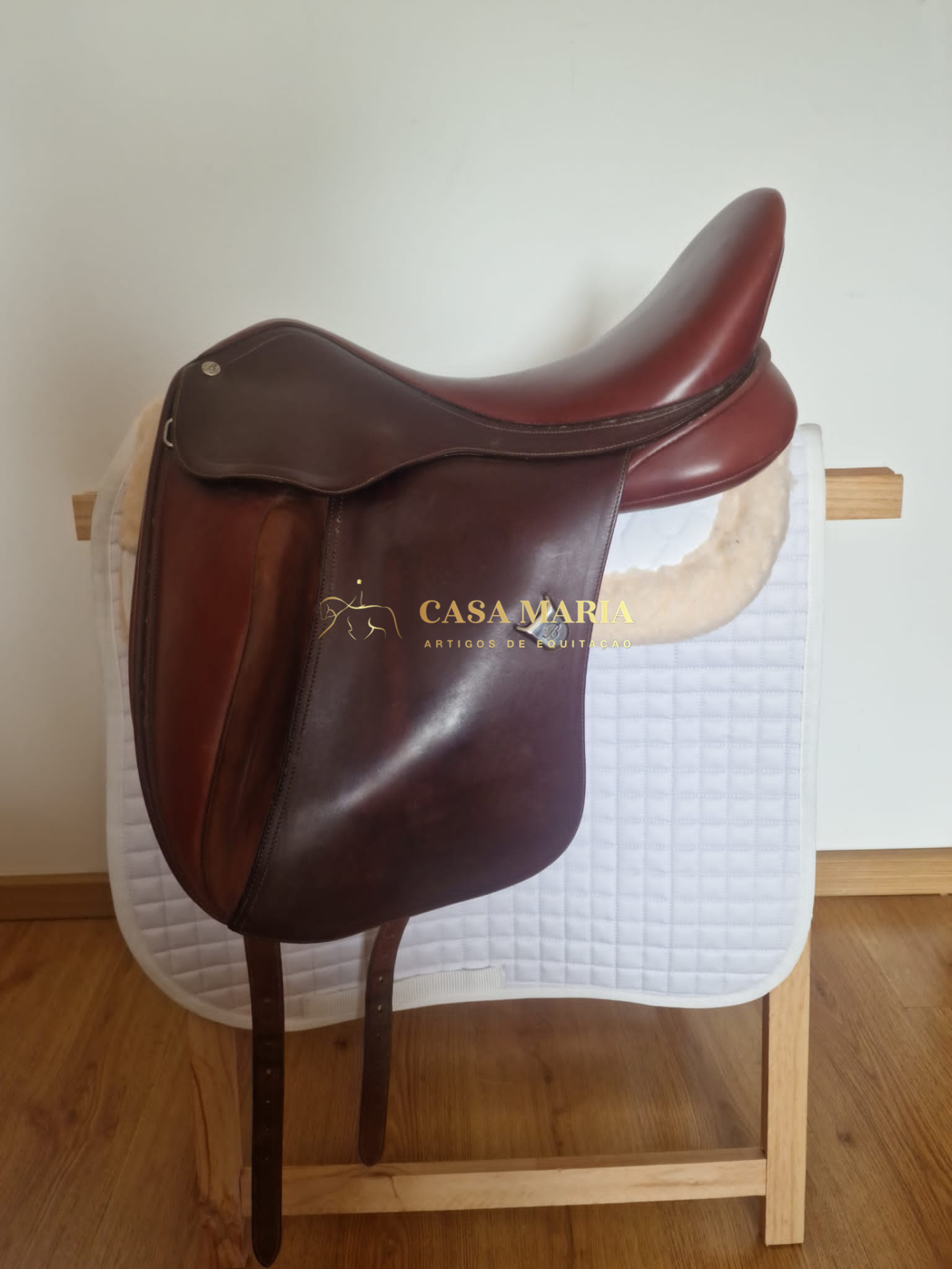 Sela de equitação em pele castanha sobre almofada branca em suporte de madeira