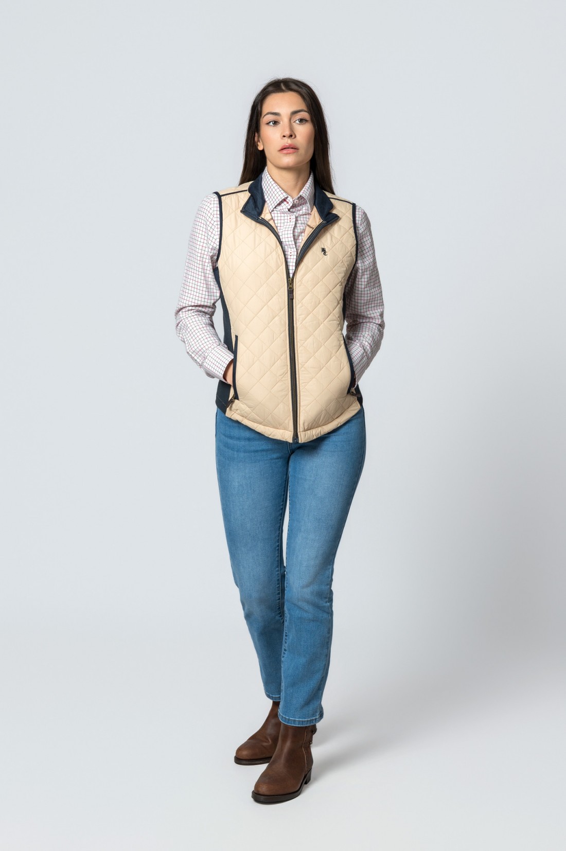 Mulher vestindo colete acolchoado bege com camisa xadrez, jeans e botins castanhos.