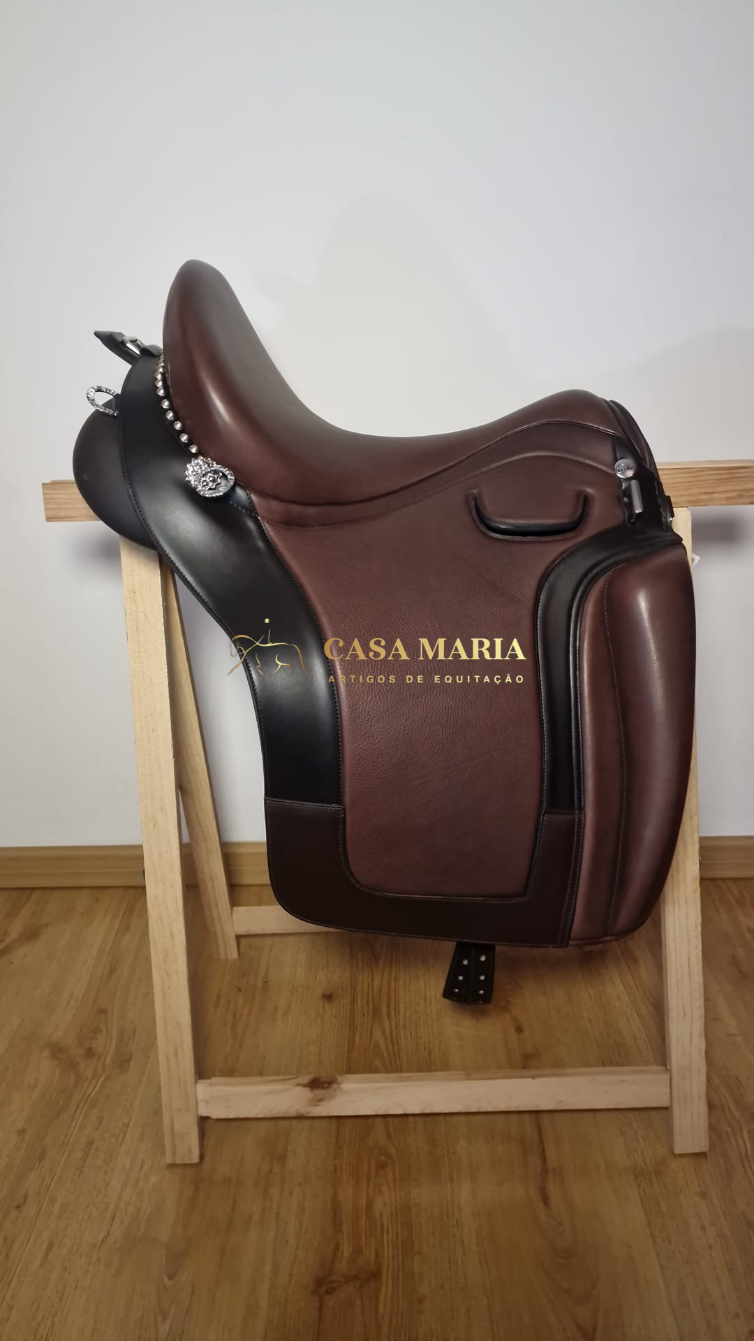 Sela de equitação em couro castanho e preto com detalhes metálicos, apoiada numa estrutura de madeira.