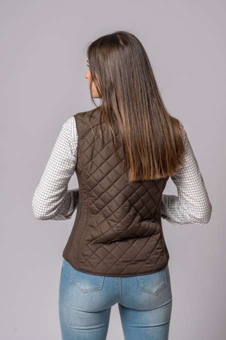 Colete acolchoado castanho escuro em padrão xadrez numa mulher de costas com camisa branca de riscas e jeans azuis claros