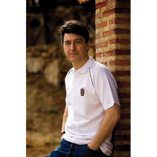 Homem com camisa polo branca e calça jeans encostado em parede de tijolos