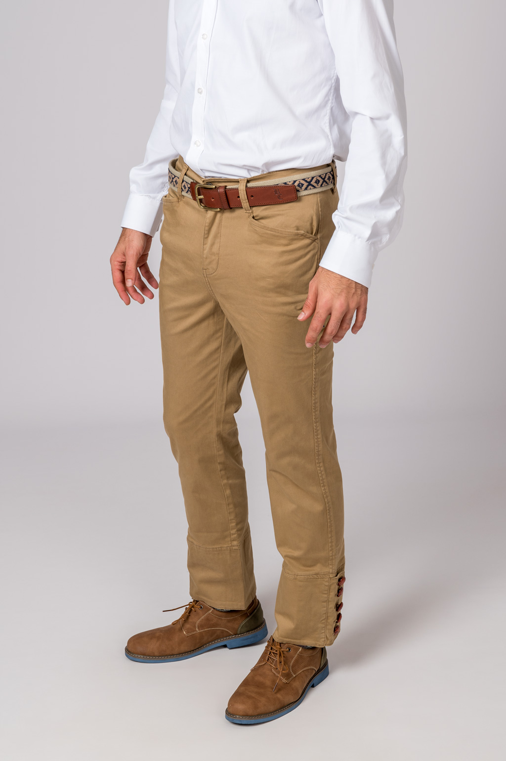 Calças bege com botões na bainha, cinto castanho com padrão, camisa branca e sapatos castanhos.