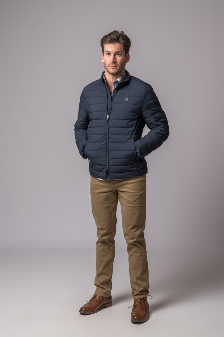 Homem com casaco azul escuro acolchoado, calças castanhas e sapatos castanhos em fundo cinzento.