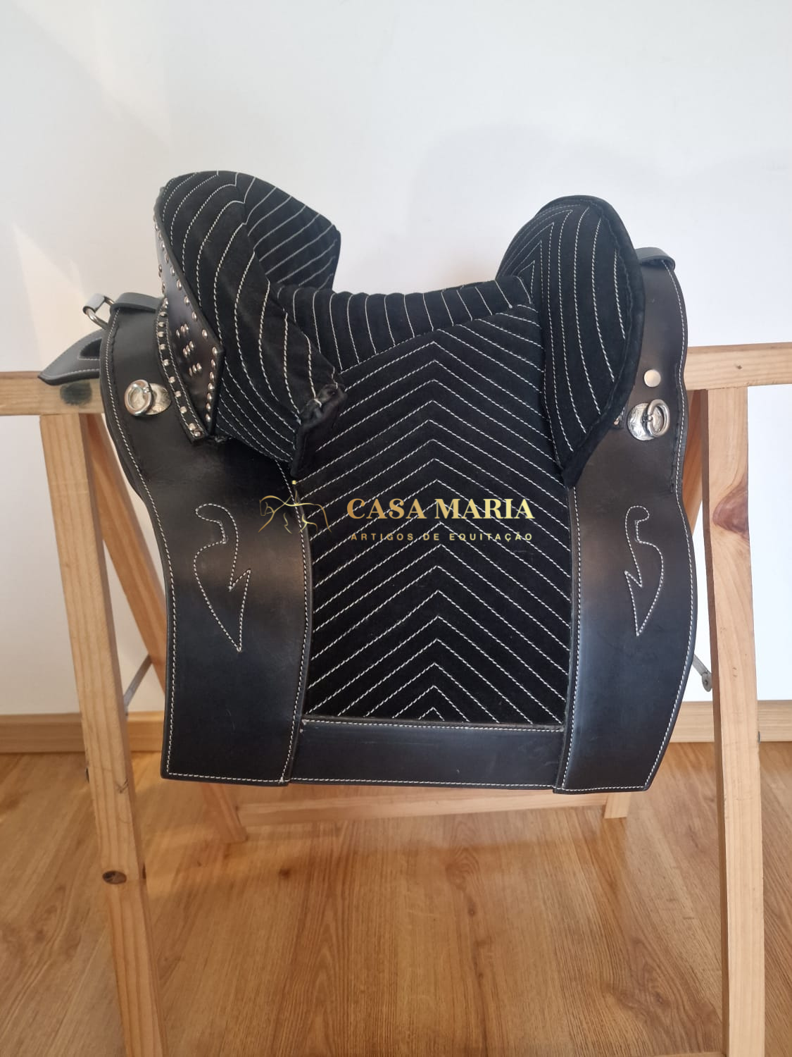 Sela de equitação preta e branca com detalhes metálicos numa estrutura de madeira