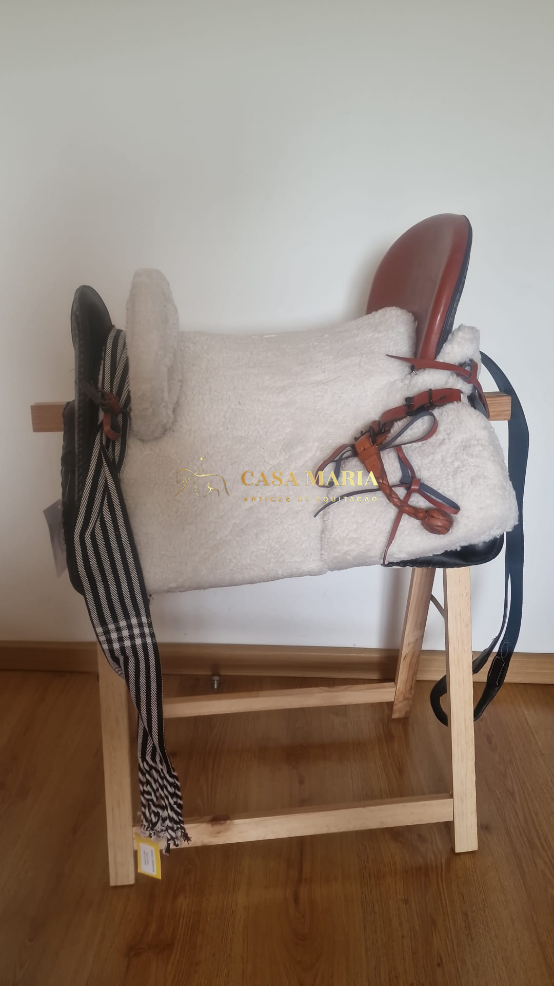 Sela de montar em couro castanho com almofadado branco e cintas riscas preto branco penduradas em suporte de madeira
