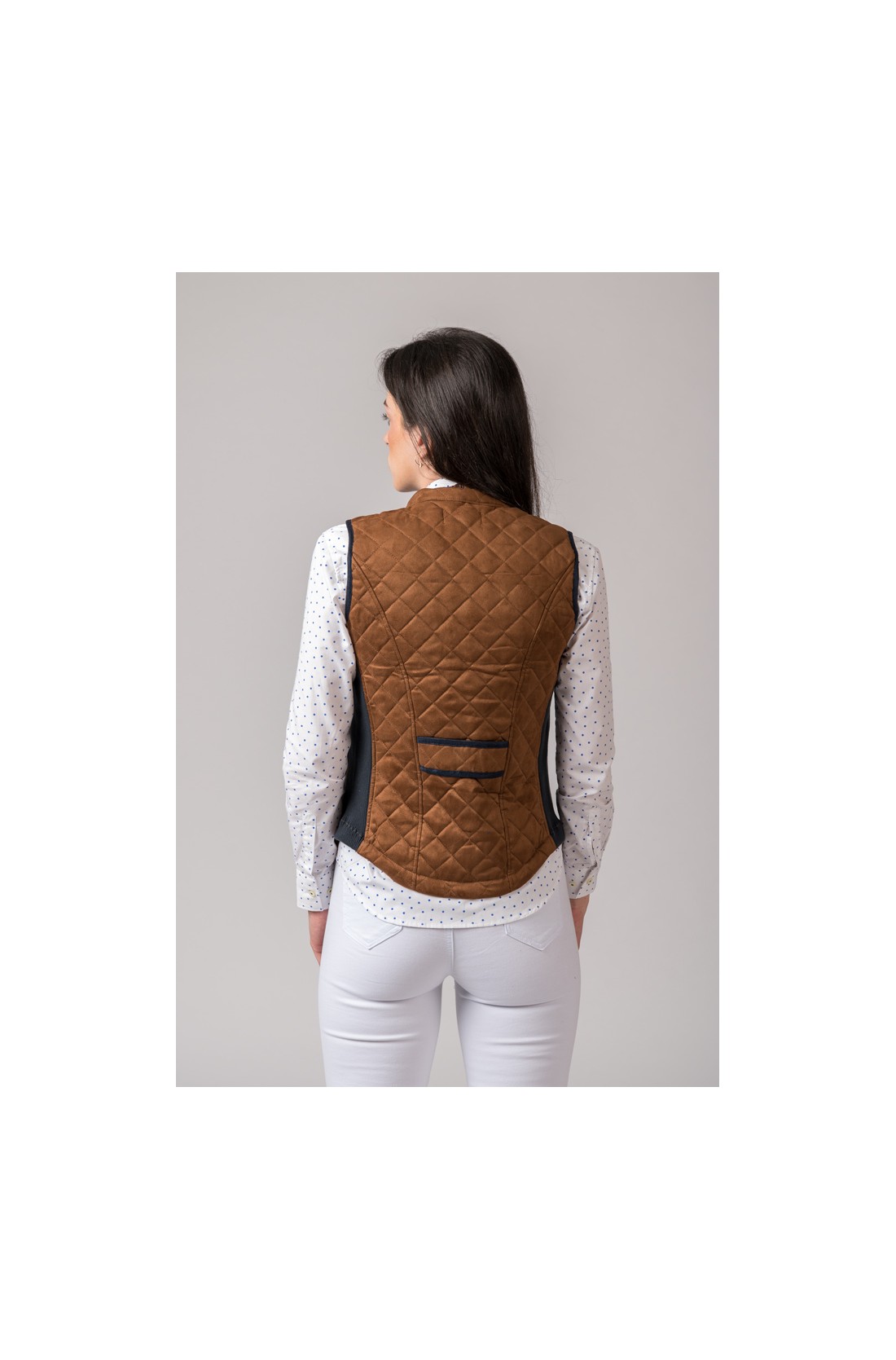 Colete feminino castanho acolchoado com bolso pequeno preto nas costas, camisa branca com padrão e calças brancas