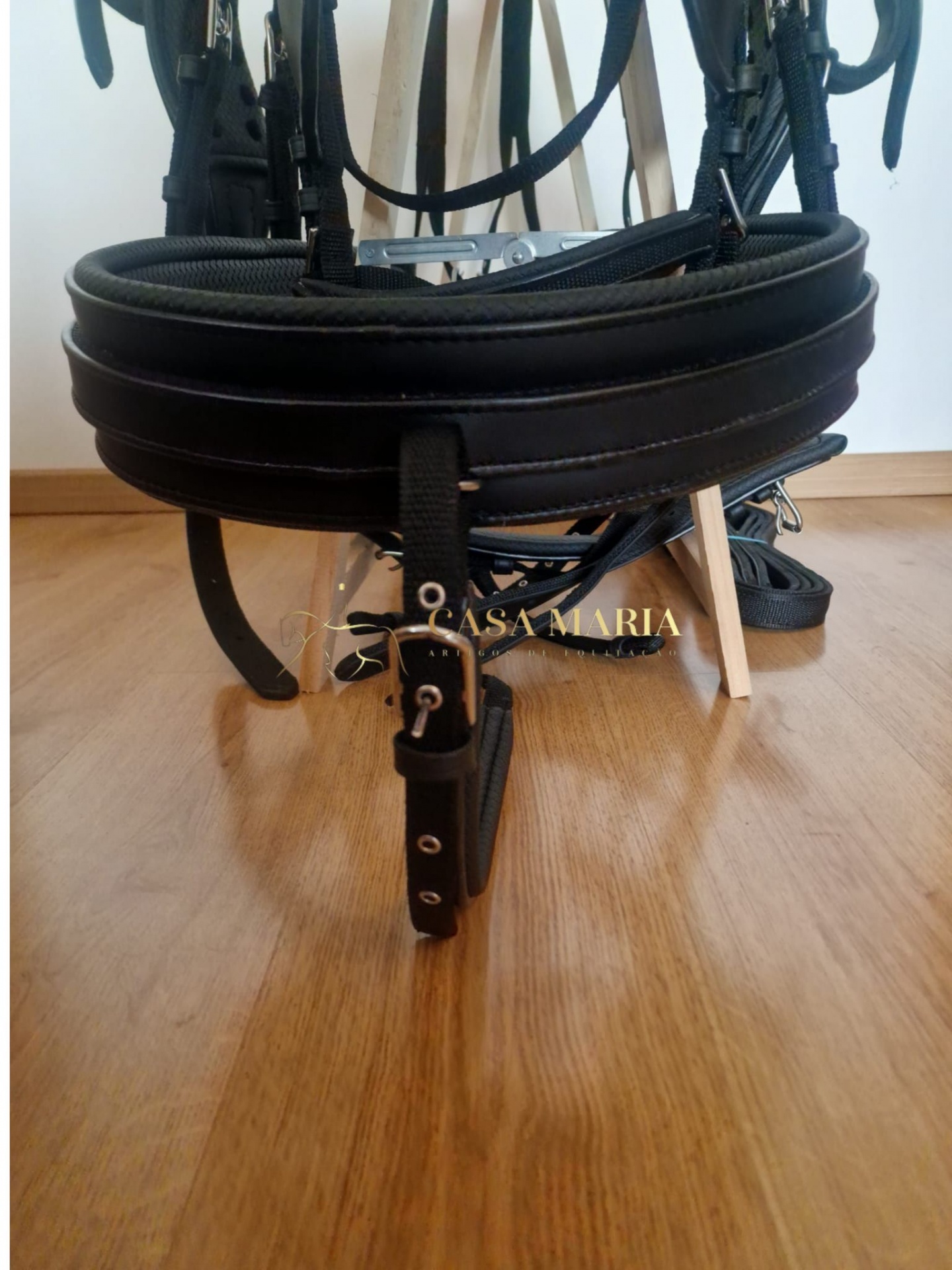 Produto de equitação de couro preto com fivelas metálicas prateadas em chão de madeira.