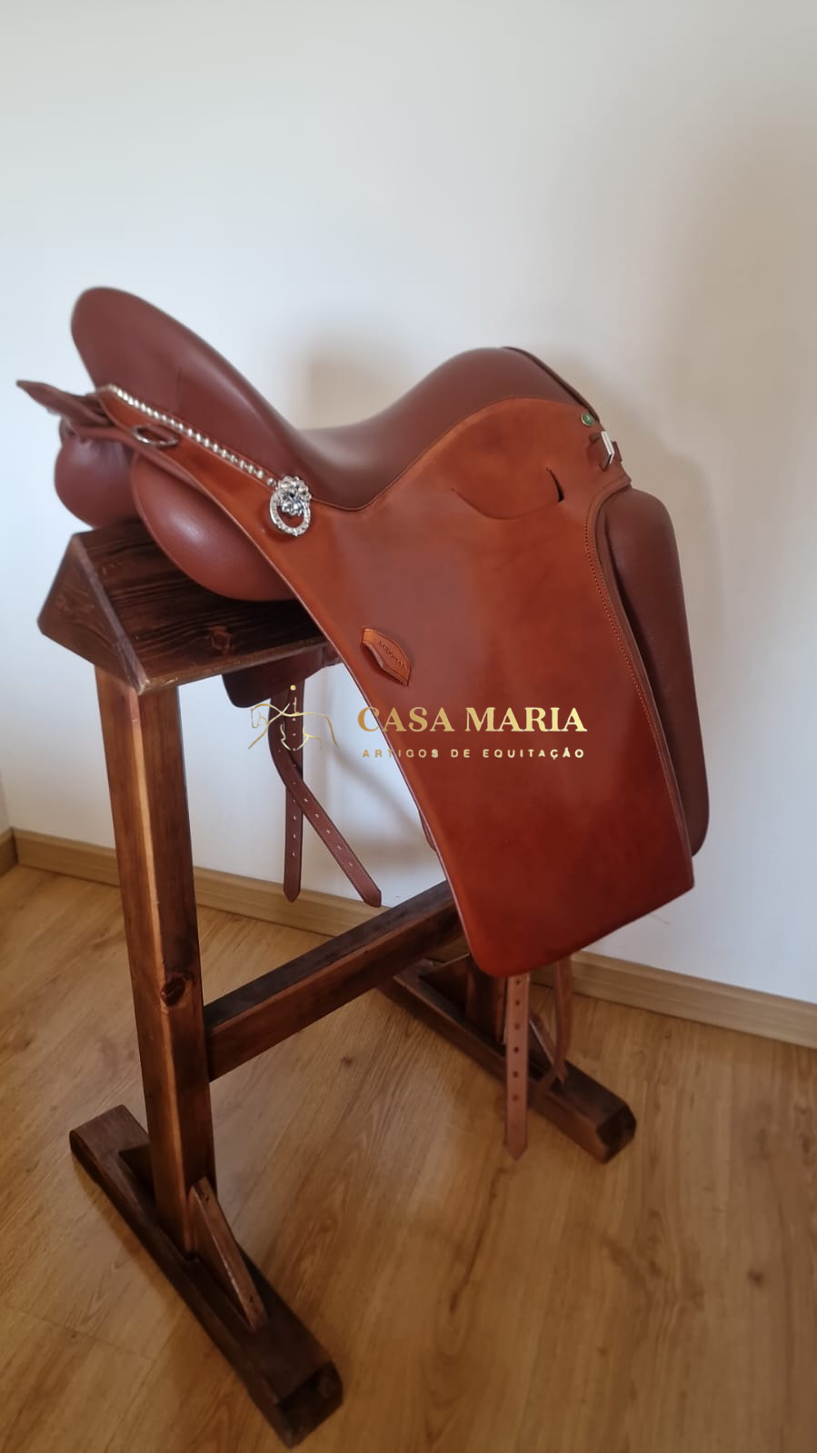 Sela de equitação castanha em suporte de madeira e texto CASA MARIA ARTIGOS DE EQUITAÇÃO