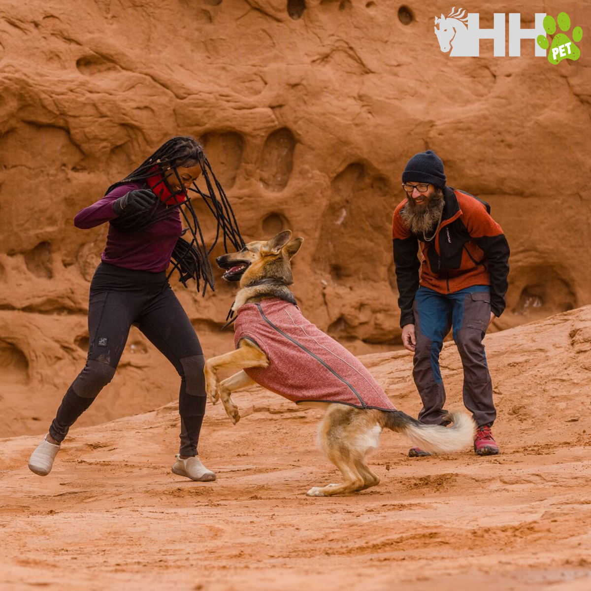 Cão pastor com casaco rosa e duas pessoas em ambiente rochoso