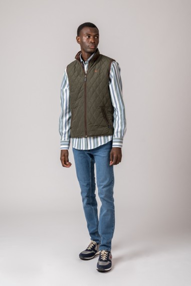 Homem com colete verde-oliva matelassê, camisa de riscas azul claras e brancas, calças de ganga e sapatilhas azul e bege