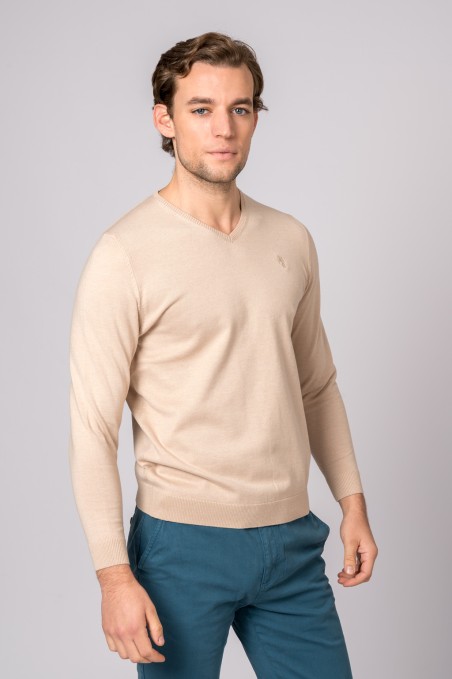 Homem com camisola de malha bege e calças azuis em fundo cinzento