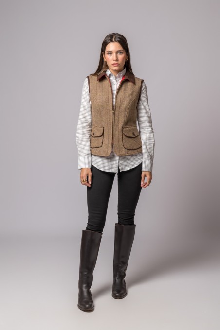 Mulher com colete castanho, camisa xadrez, calças pretas e botas pretas sobre fundo cinzento