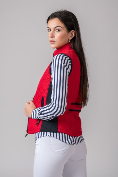 Mulher de perfil com colete vermelho acolchoado, camisa às riscas azul e branca e calças brancas