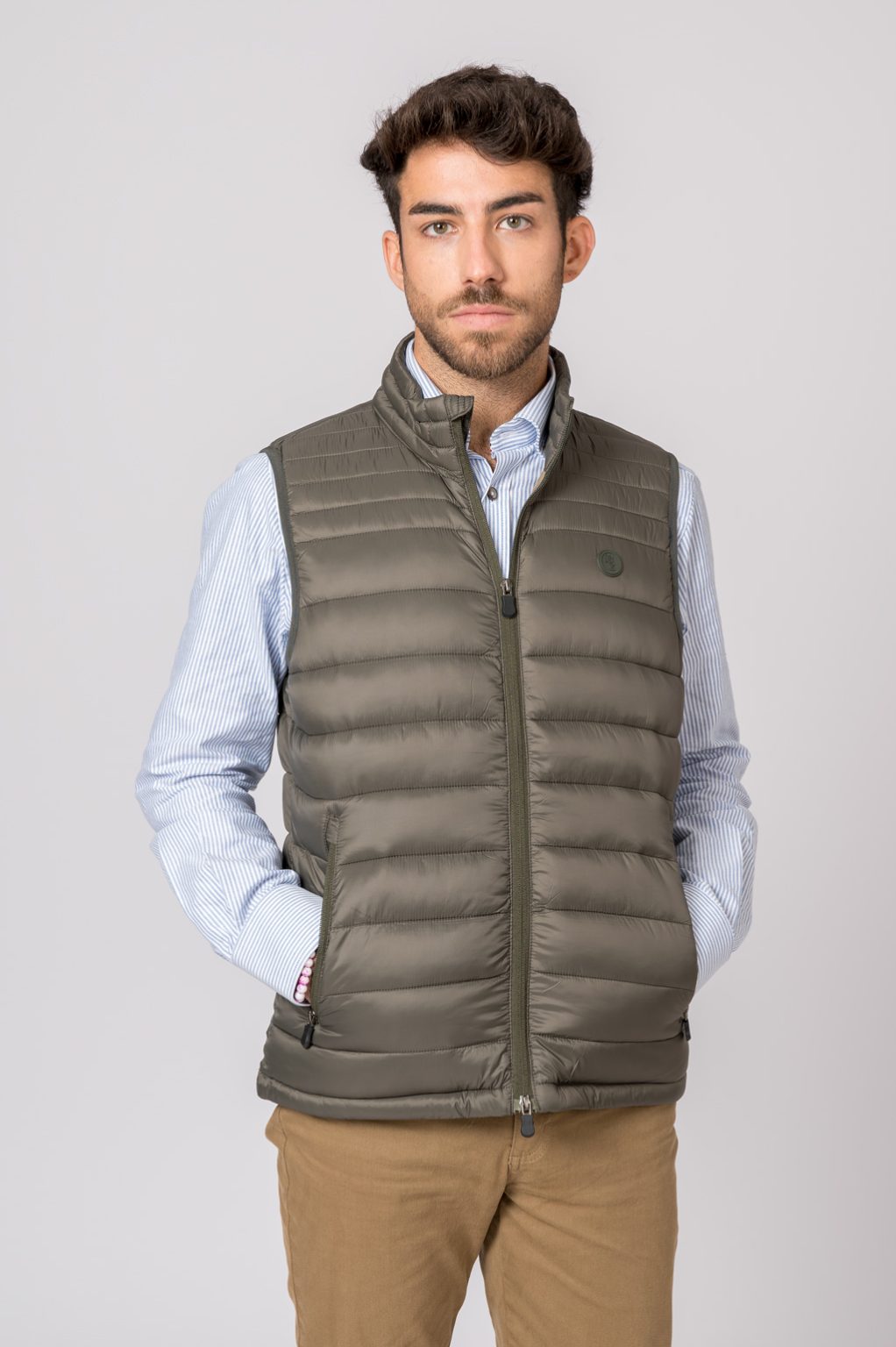 Colete acolchoado verde-oliva com fecho e bolso, usado com camisa azul clara e calças castanhas
