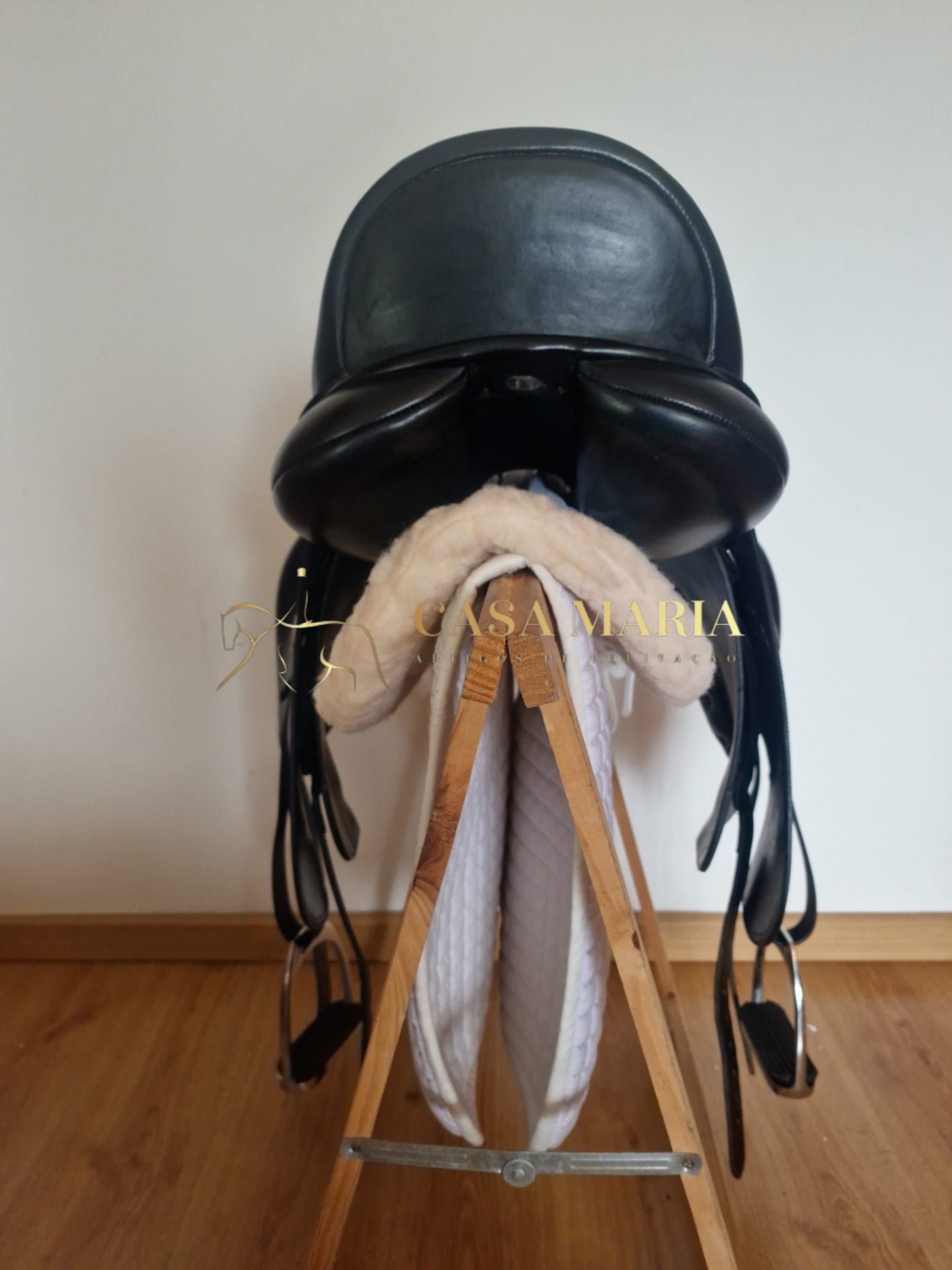Sela de equitação preta em couro com manta e barrigueira brancas sobre suporte de madeira