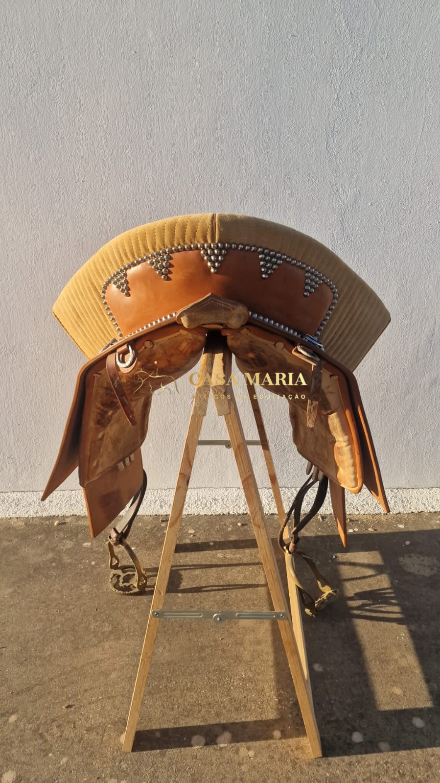 Selim de cavalo castanho claro e bege com decoração metálica em suporte de madeira