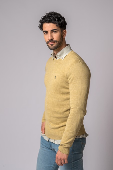 Homem com camisola amarelo claro, camisa bege e calças de ganga azuis em fundo cinzento.