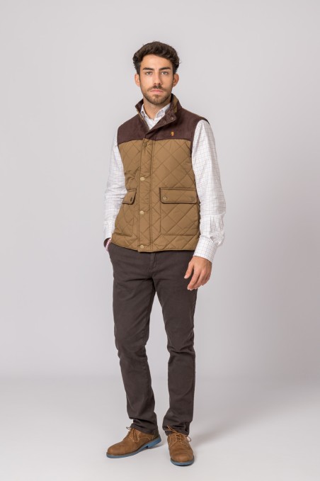 Homem com colete acolchoado castanho, camisa xadrez branca, calças castanhas e sapatos castanhos sobre fundo cinza claro