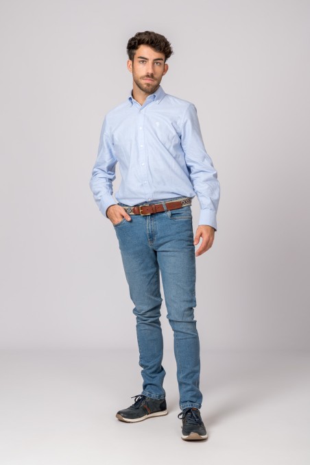 Homem com camisa azul clara, jeans azul, cinto castanho e sapatos pretos num fundo cinzento.
