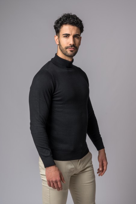 Homem com camisola preta de gola alta e calças bege, fundo cinzento.