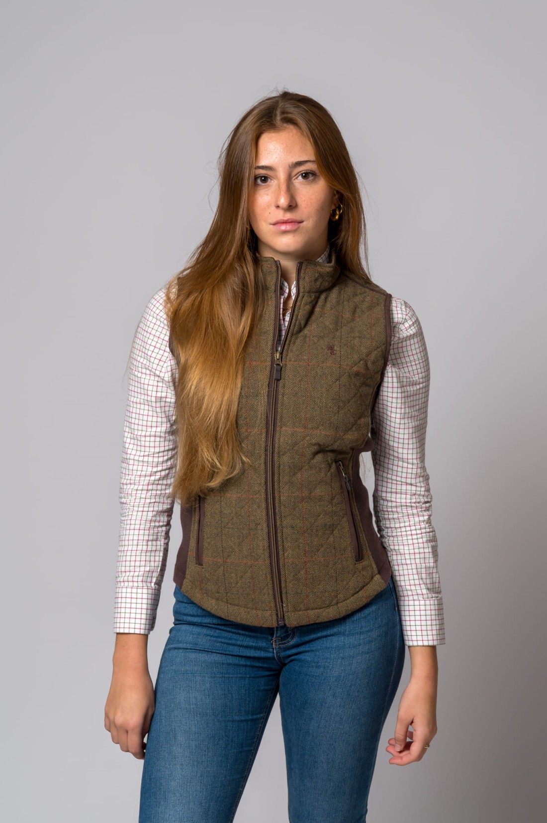 Mulher com colete castanho e camisa xadrez em fundo cinzento